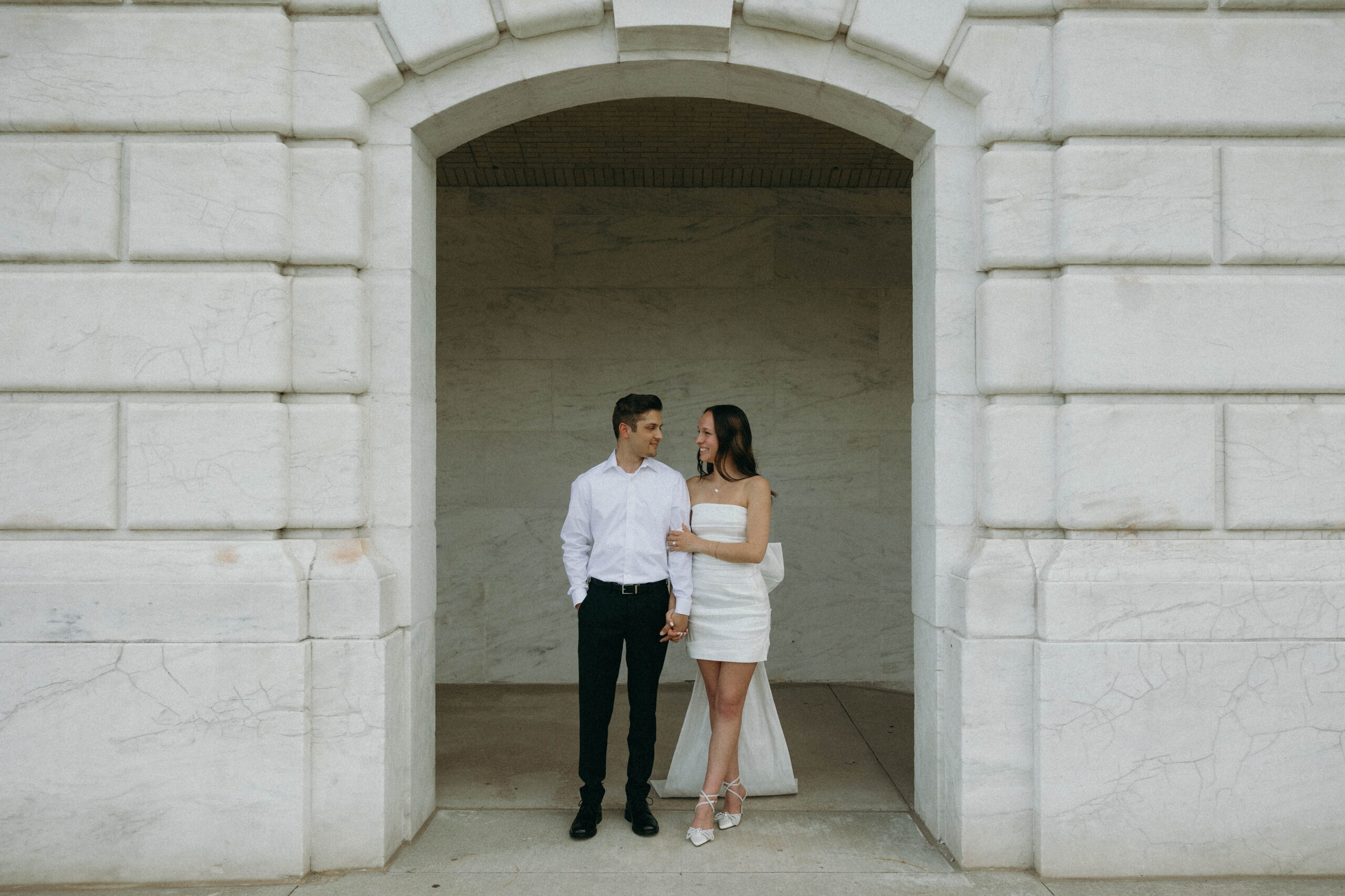 future bride and groom posing together during engagement photo shoot, bride-to-be is wearing a short white strapless dress with a big bow in the back; groom is wearing black slacks and black dress shoes with a timeless, long sleeve button down shirt. they're standing in front of a marble building exterior in detroit, Michigan