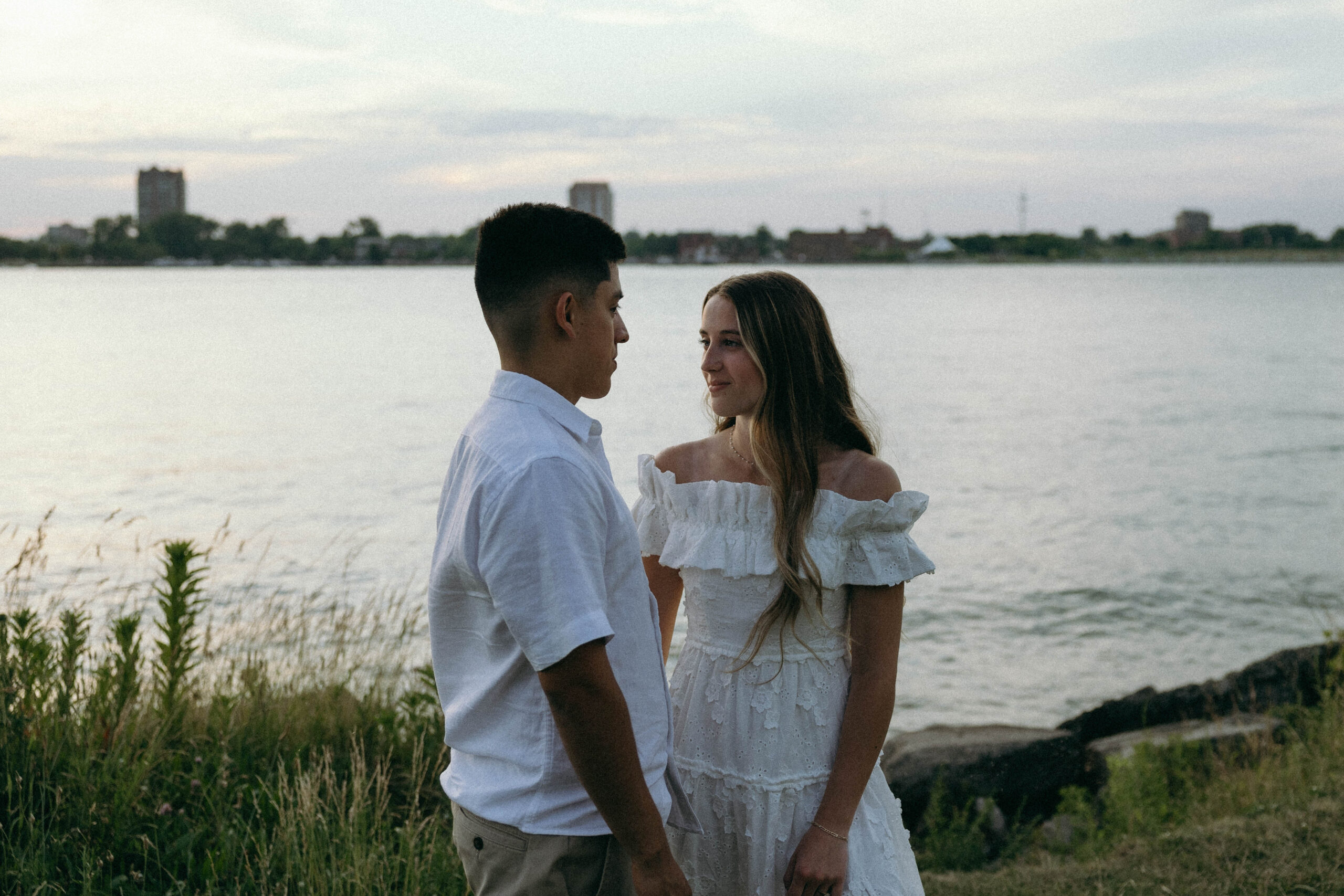 A couple posing in the evening time for engagement photos by a serene lake, surrounded by lush greenery and a blue sky.