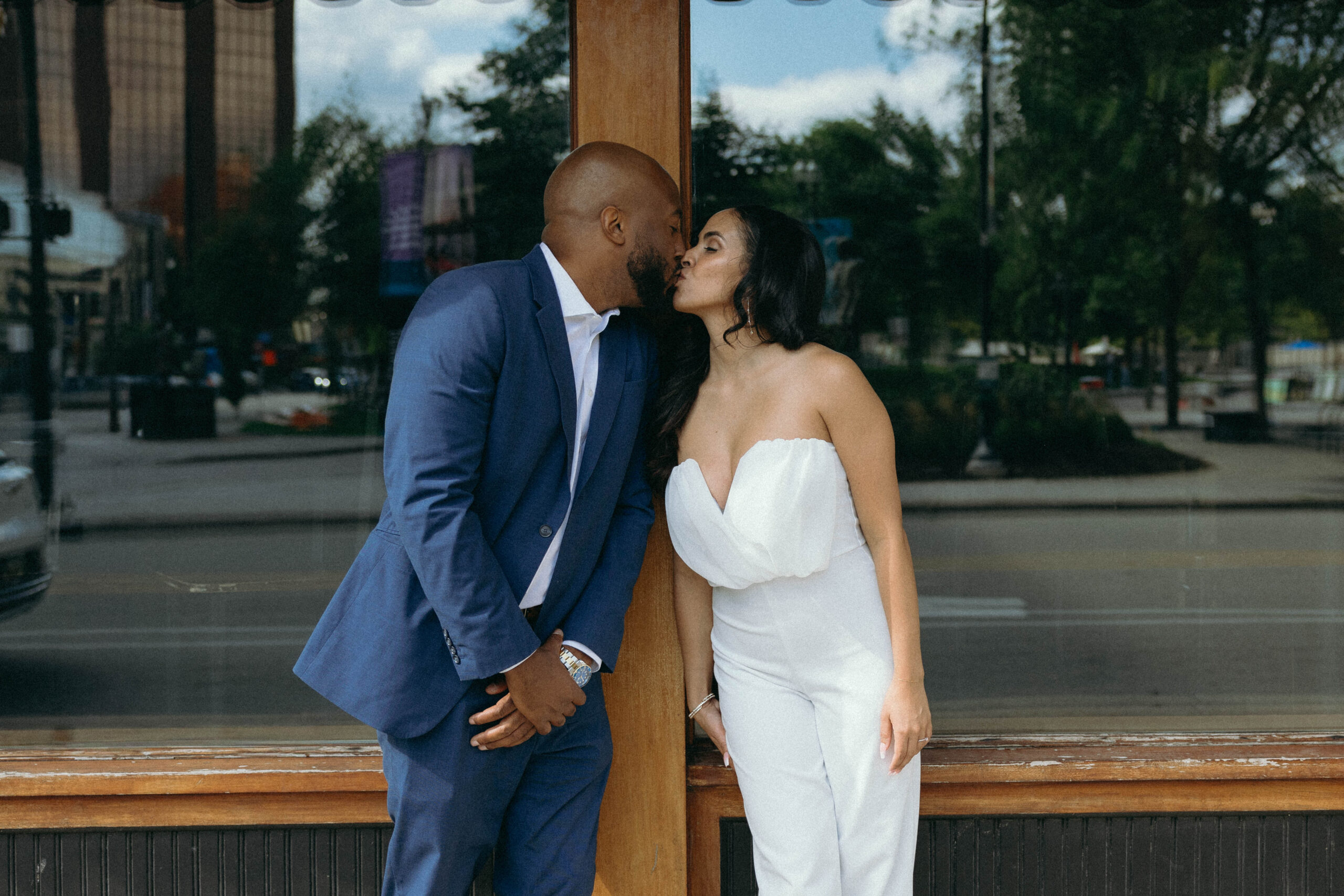 Man in blue suit kissing a woman in a white jumpsuit in front of a brick building with glass windows during their engagement photo session.