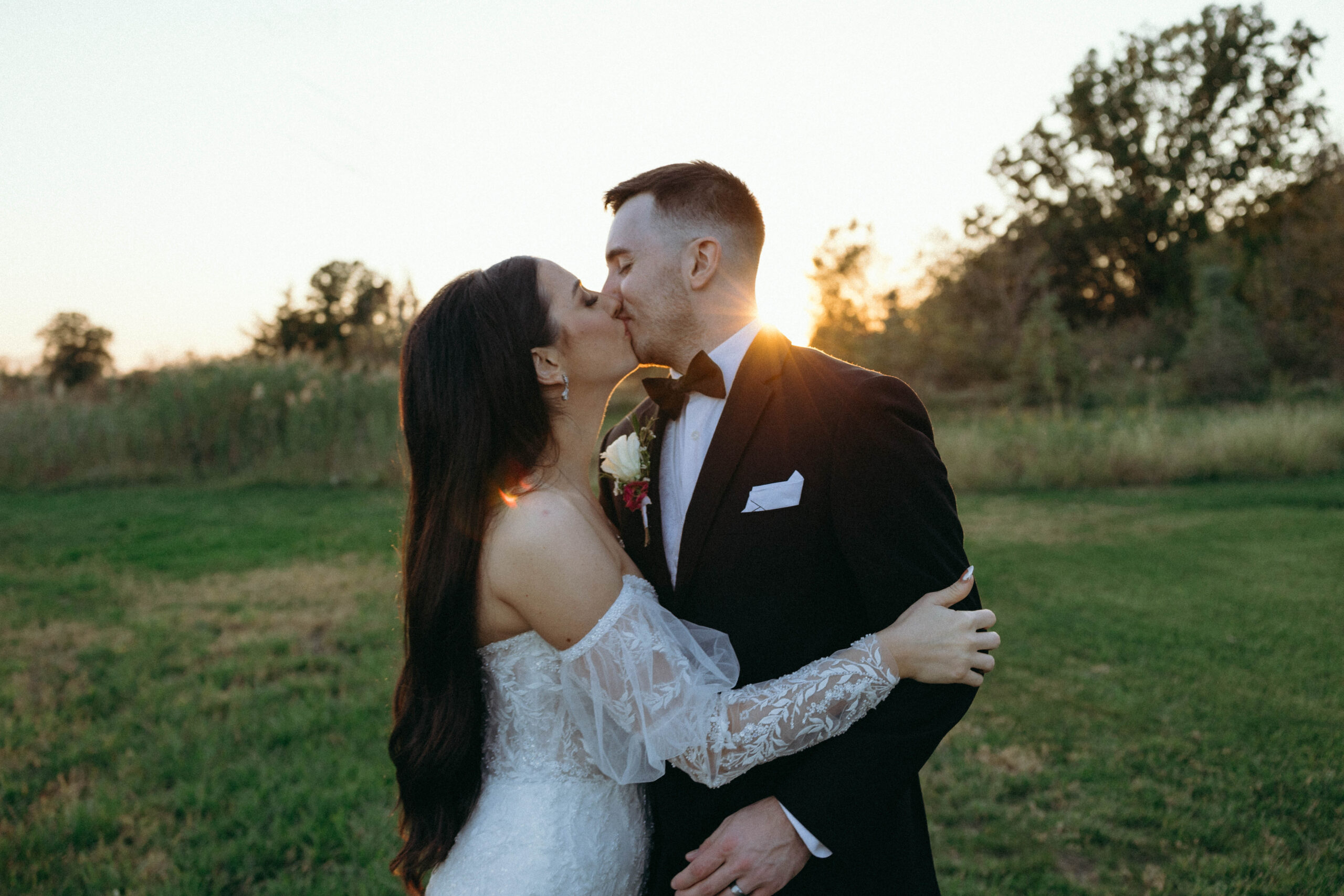 bride and groom on wedding day, kissing in a green field at sunset