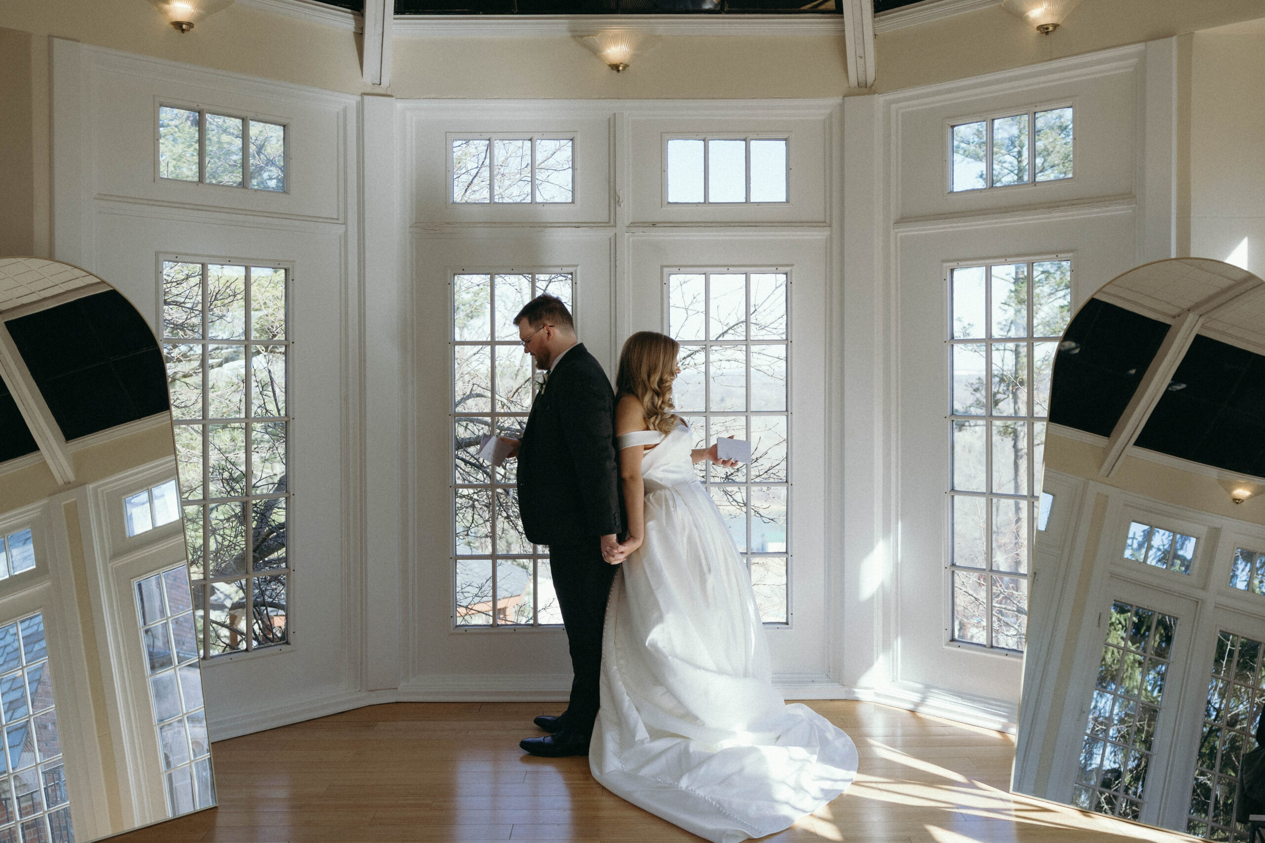 A bride and groom stand together in front of large windows, showcasing their elegant wedding attire and joyful expressions while exchanging private vows before the ceremony.