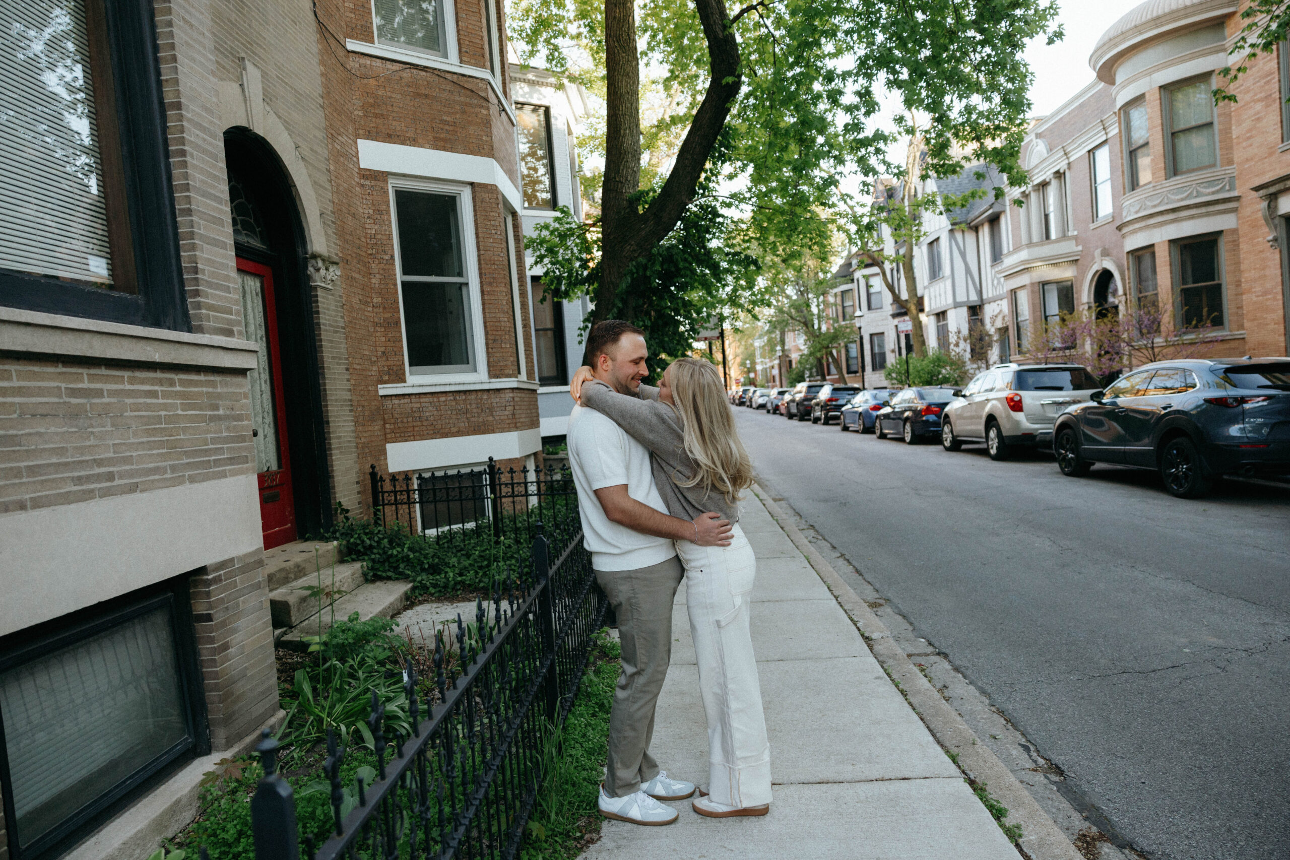 An engaged couple embraces on the sidewalk, surrounded by a row of charming houses in the background.