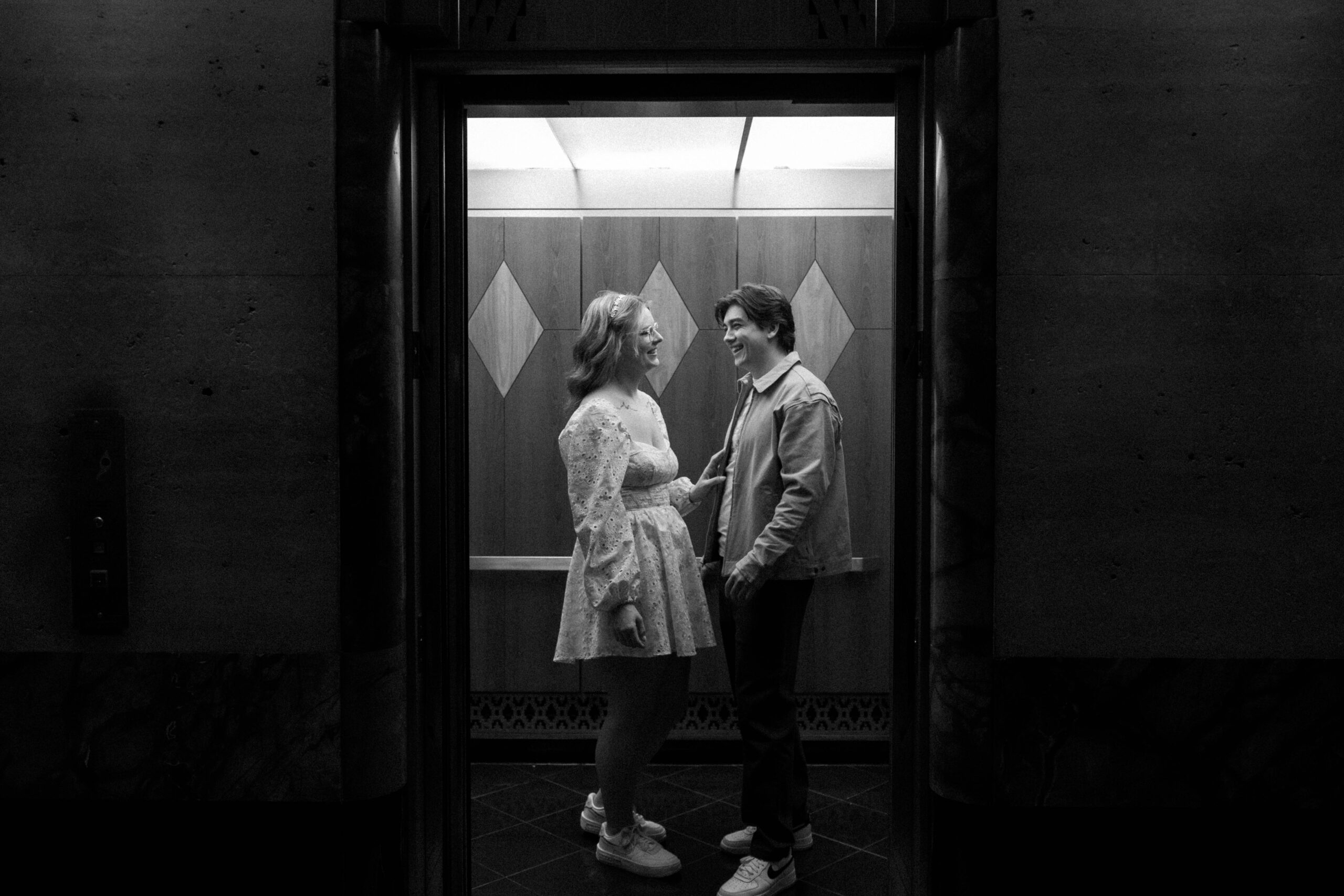 A couple stands in an open elevator, smiling and looking at each other. The setting is in black and white, creating an intimate, nostalgic mood.