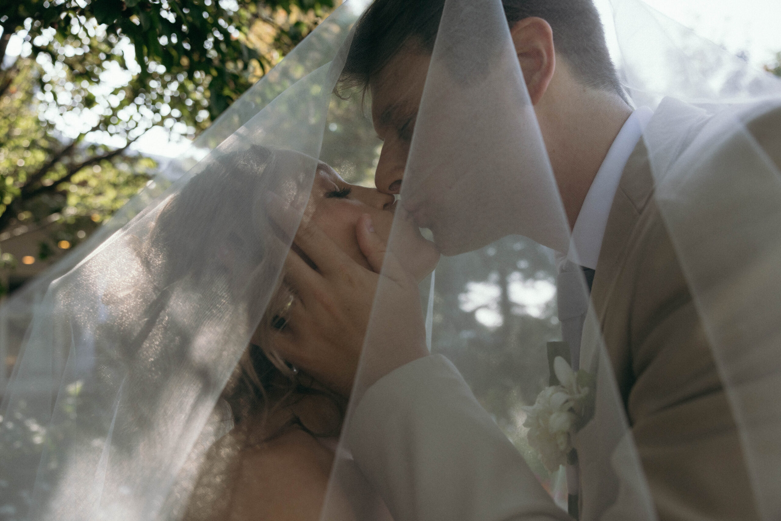 A couple under a veil shares an intimate moment outdoors, with soft natural light filtering through the trees on the wedding day.