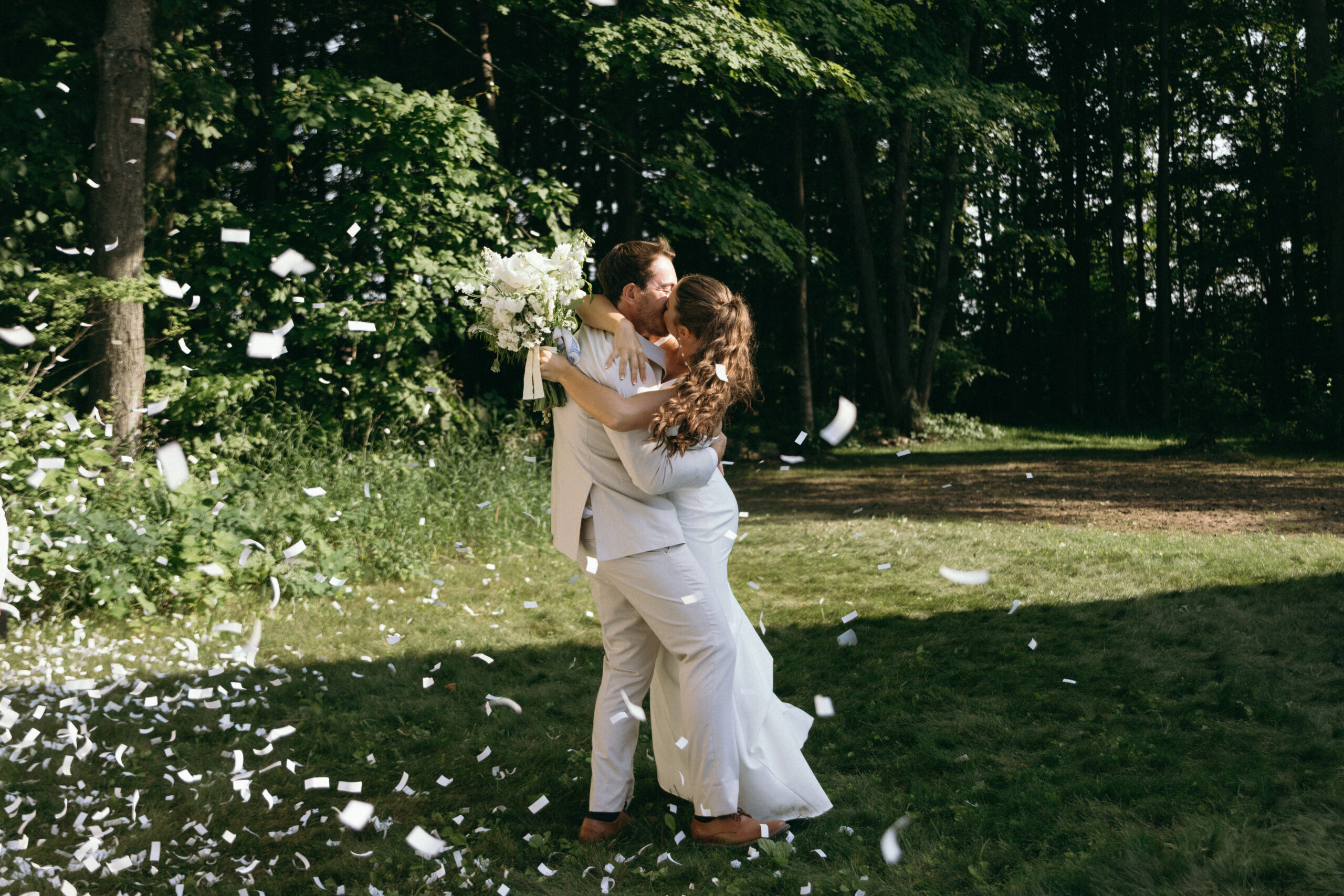 A couple in wedding attire embraces and kisses outdoors as flower petals or confetti fall around them, surrounded by trees and grass.