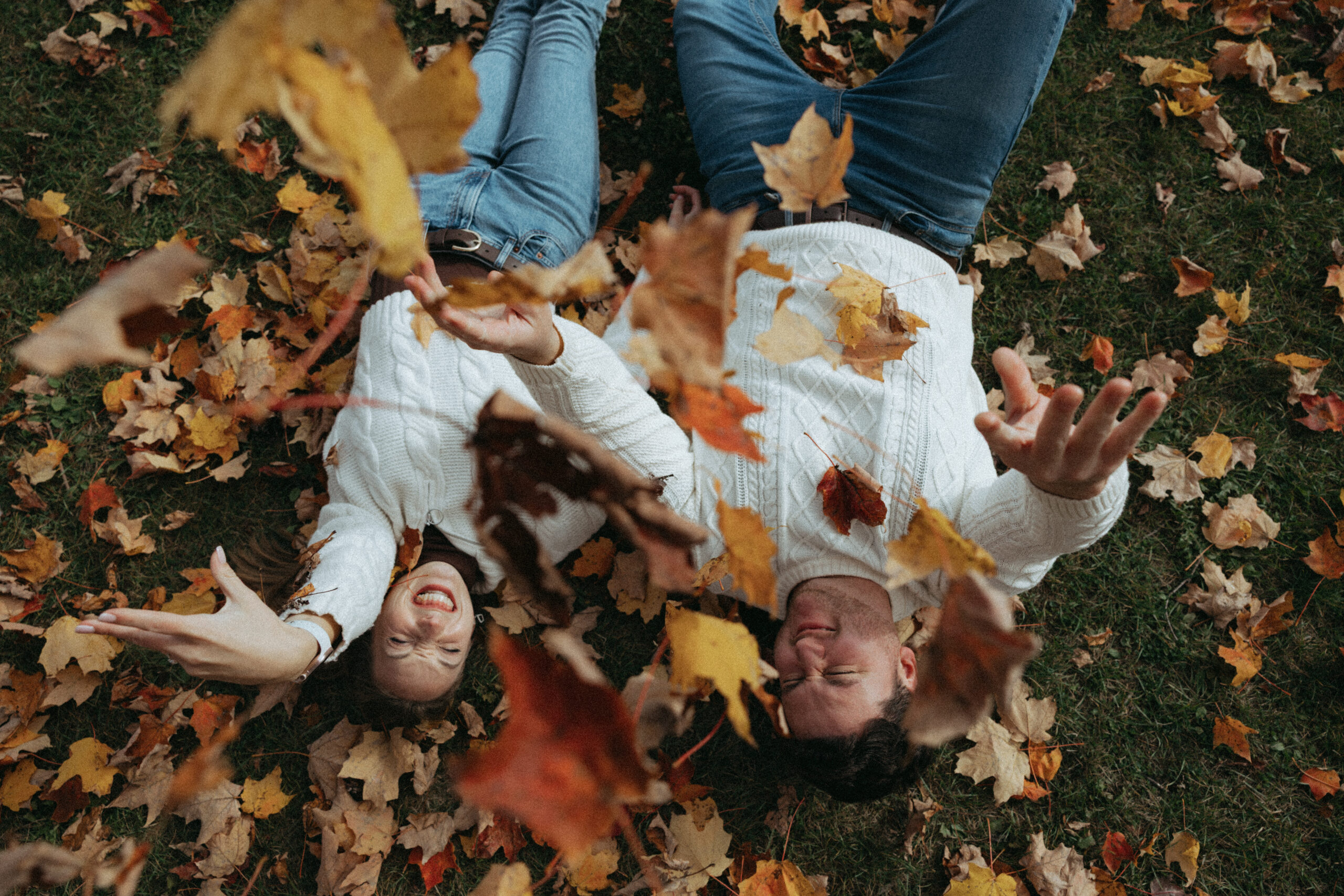 A young couple laying in the grass throwing fall leaves in the air during their engagement photo session.