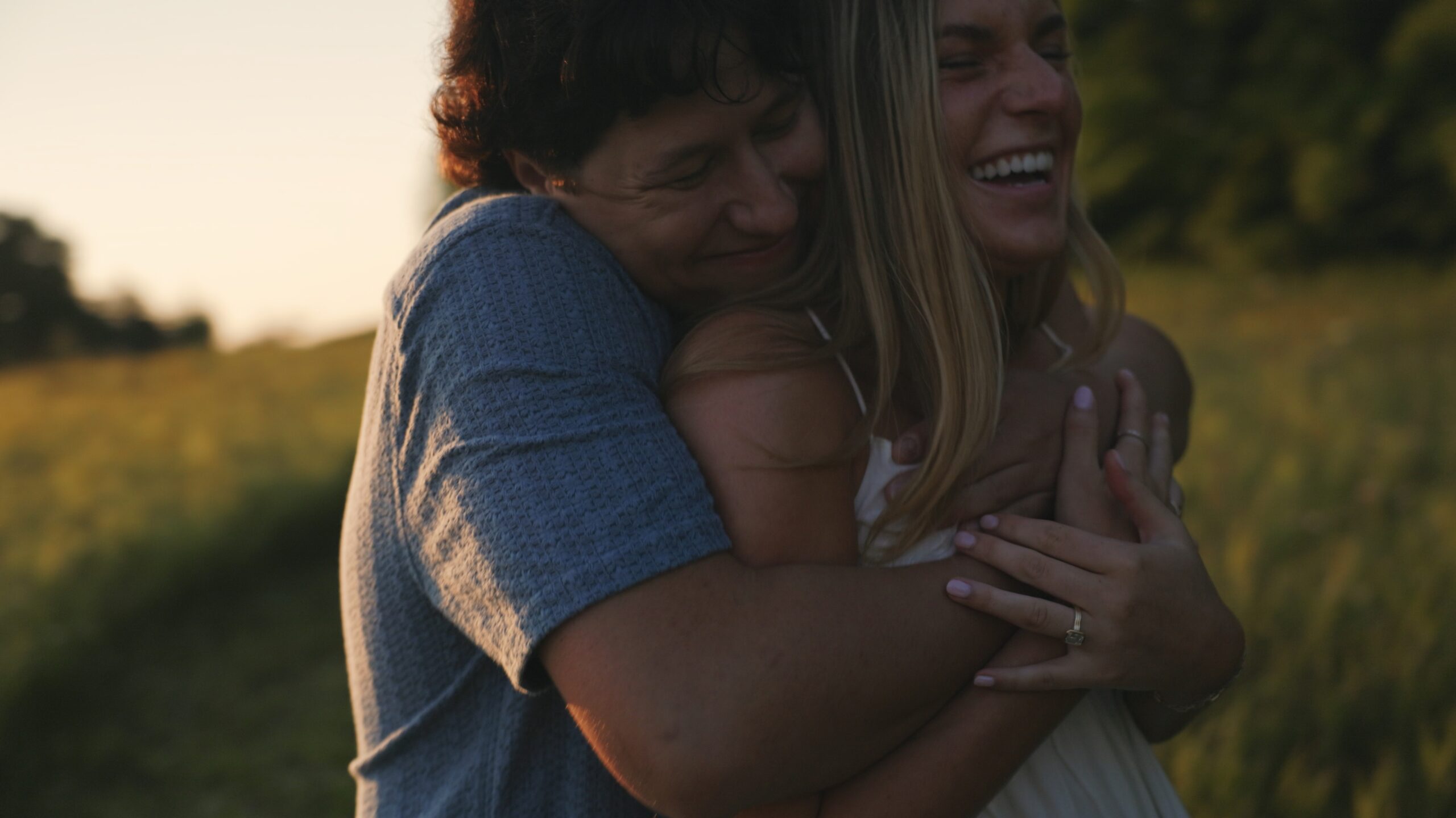 A woman embraces her fiancé in a sunny field, surrounded by tall grass and wildflowers, conveying warmth and love.