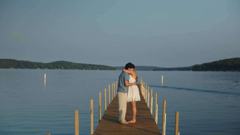 A man and woman stand together on a wooden dock, overlooking a calm body of water under a clear blue sky.  