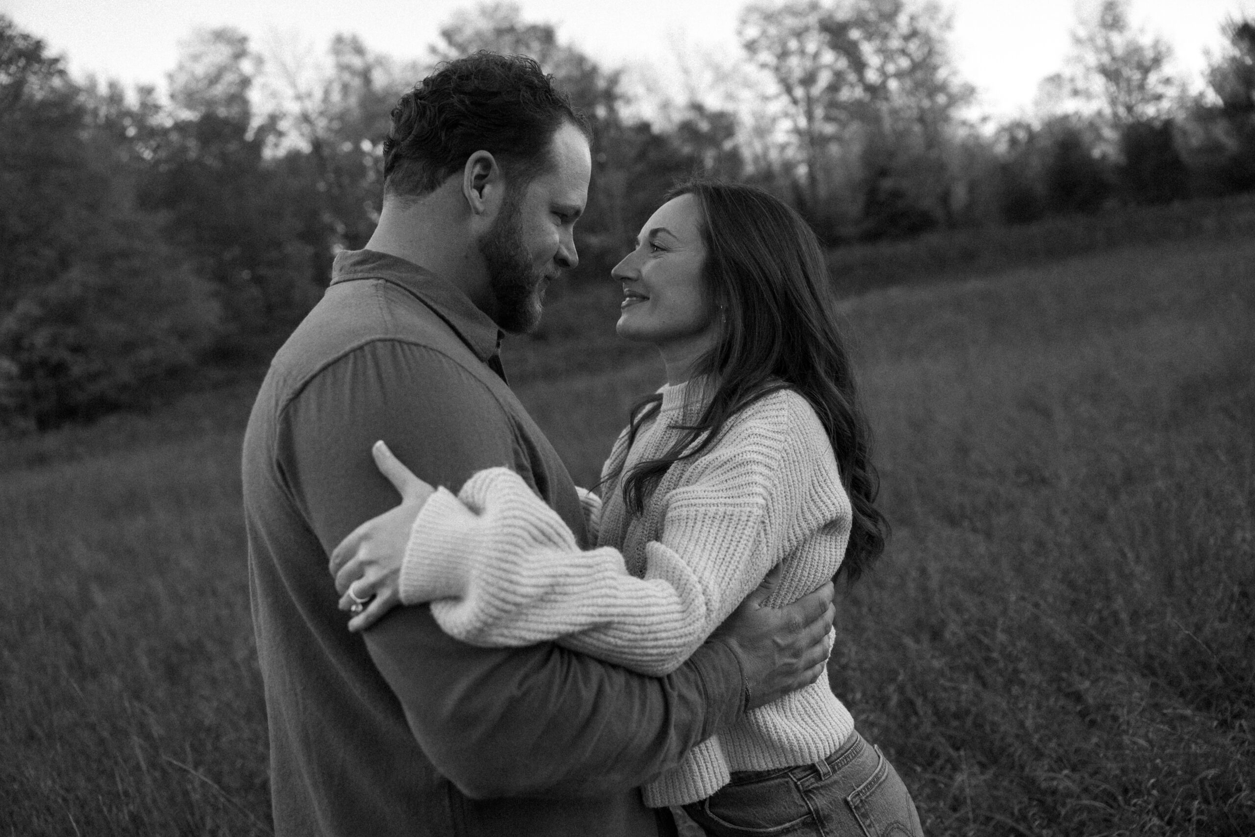 A black and white photo of an engaged couple walks around field during the fall season, capturing this season of their love story.