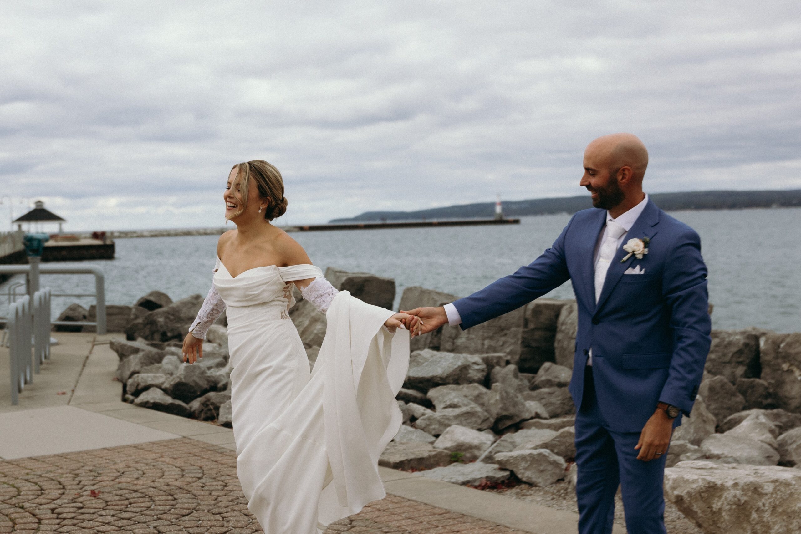 The bride and groom walk together near the water, enjoying a scenic view of boats on the water in northern michigan.